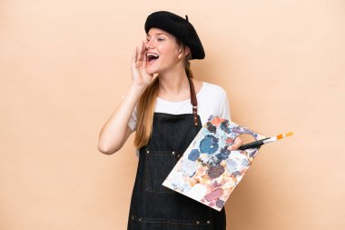 Beyaz tenli genç bir kadın bej arka planda izole edilmiş bir palet tutuyor ve ağzı sonuna kadar açık bağırıyor.