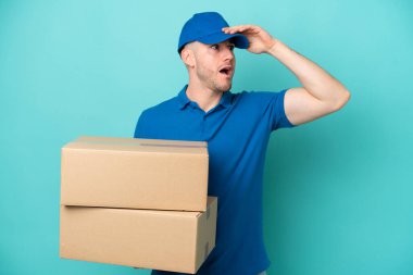 Teslimat yapan beyaz adam mavi arka planda izole edilmiş. Yan tarafa bakarken sürpriz jest yapıyor.