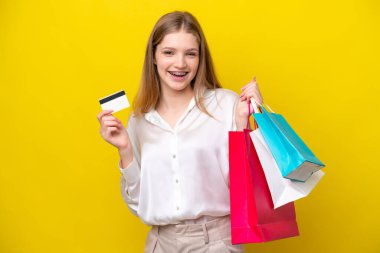 Sarı arka planda izole edilmiş genç bir Rus kız elinde alışveriş çantaları ve bir kredi kartı tutuyor.