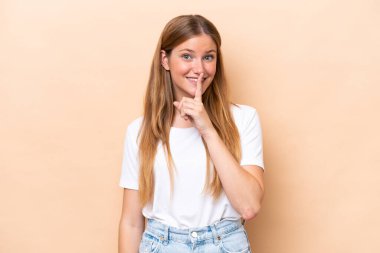 Beyazlı genç bir kadın bej arka planda izole edilmiş. Parmağını ağzına koyduğunda sessizlik işareti gösteriyor.