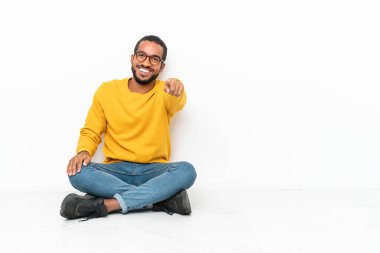 Genç Ekvadorlu, beyaz duvarda izole bir şekilde yerde oturuyor. Kendini güvende hisseden bir ifadeyle seni işaret ediyor.