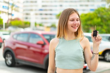 Açık havada arabanın anahtarlarını tutan genç sarışın kadın mutlu bir ifadeyle.