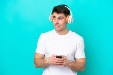 Kafkasyalı genç adam mavi arka planda izole edilmiş. Cep telefonuyla müzik dinliyor ve düşünüyor.
