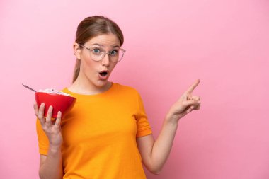 Beyaz bir kadın elinde bir kase mısır gevreği tutuyor. Pembe arka planda izole edilmiş. Şaşırmış ve işaret ediyor.