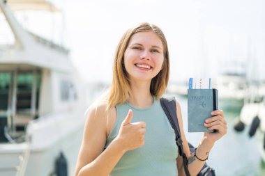 Açık havada pasaportu olan genç sarışın bir kadın. Başparmakları havada. Çünkü iyi bir şey oldu.