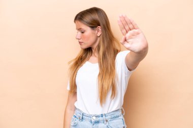 Beyaz tenli genç bir kadın bej arka planda izole edilmiş. Durma hareketi yapıyor ve hayal kırıklığına uğruyor.