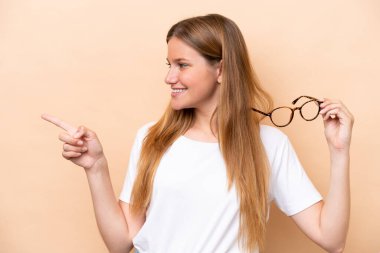 Bej arka planda gözlüklü güzel sarışın kadın bir ürünü sunmak için yan tarafı işaret ediyor.