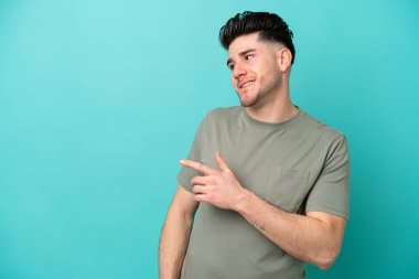 Mavi arka planda izole edilmiş genç beyaz bir adam parmağını yana doğru uzattı ve bir ürün sundu.