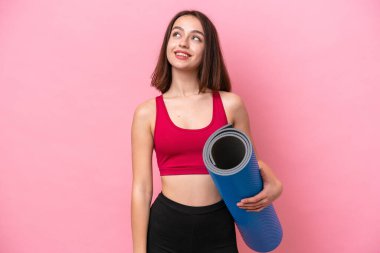 Young sport Ukrainian woman going to yoga classes while holding a mat isolated on pink background thinking an idea while looking up