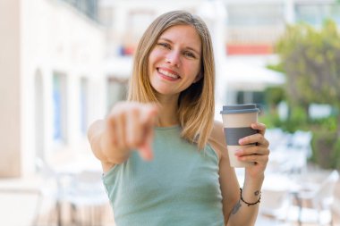 Açık havada elinde kahve tutan genç sarışın kadın kendinden emin bir ifadeyle seni işaret ediyor.