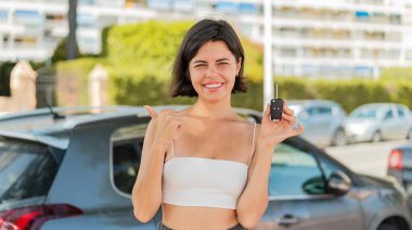 Genç, güzel bir Bulgar kadın dışarıda araba anahtarı tutuyor ve bir ürün sunmak için kenarı işaret ediyor