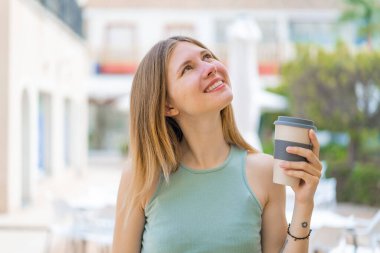 Sarışın genç bir kadın açık havada kahvesini yudumlarken gülümsüyor.