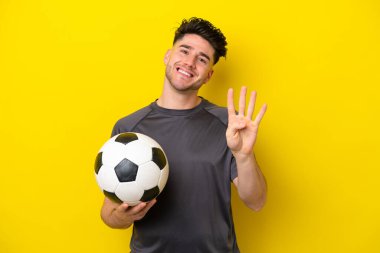 Yakışıklı genç futbolcu sarı arka planda izole edilmiş mutlu ve parmaklarıyla dörde kadar sayıyor.