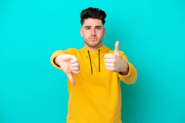 Kafkasyalı genç adam mavi arka planda izole edilmiş, iyiye-kötüye işaret yapıyor. Evet ya da hayır arasında kararsız