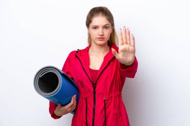 Genç bir kız yoga derslerine giderken elinde beyaz bir hasırla durup jest yapıyor.