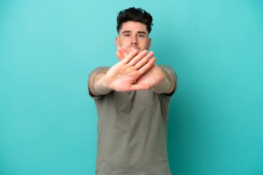 Kafkasyalı genç adam mavi arka planda izole edildi. Bir hareketi durdurmak için eliyle dur hareketi yapıyor.
