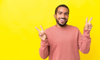 Sarı arka planda izole edilmiş genç latin adam iki eliyle zafer işareti gösteriyor.