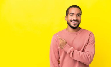 Sarı arka planda izole edilmiş genç latin adam bir ürün sunmak için kenarı işaret ediyor.