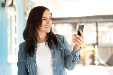 Orta yaşlı bir kadın açık havada mutlu bir ifadeyle cep telefonu kullanıyor.