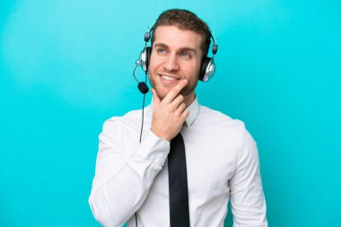 Tele pazarlamacı beyaz bir adam mavi arka planda izole edilmiş bir kulaklıkla çalışıyor. Yukarı bakarken bir fikir düşünüyor.