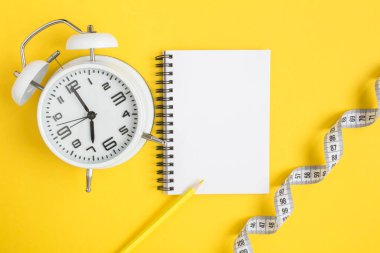 Açık not defterinin üst görünümü, beyaz alarm saati ve sarı arkaplanda beyaz santimetre