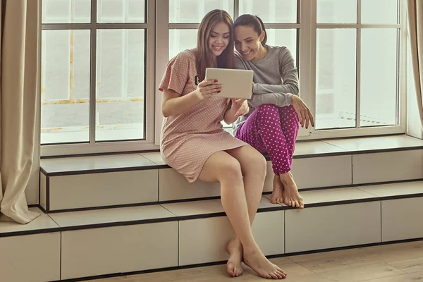 Duas mulheres em roupas de dormir estão sentadas no peitoril da janela e assistindo vídeo engraçado na tela do tablet pc. — Fotografia de Stock