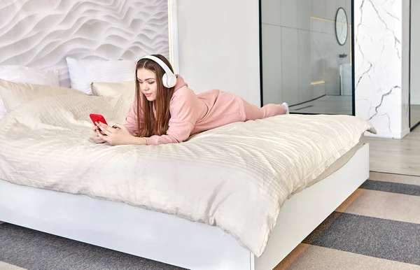 Young woman 20 years old lies in bed on her stomach and listens to music through headphones. — Stock Fotó