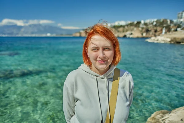 Rudowłosa Europejka przed turkusową wodą w zatoce Antalya w chłodny słoneczny dzień wczesną wiosną. — Zdjęcie stockowe