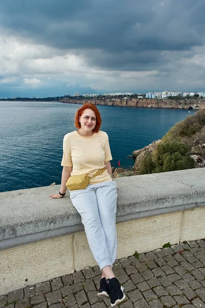 Young smiling redhead Caucasian woman posing for photographer while sitting on fence of observation point overlooking sea in Antalya. — Stock Photo, Image