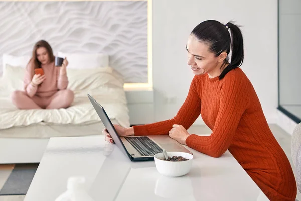 Bureau à domicile pour femme travaillant à la maison avec ordinateur portable à table et sa fille adolescente en arrière-plan. — Photo