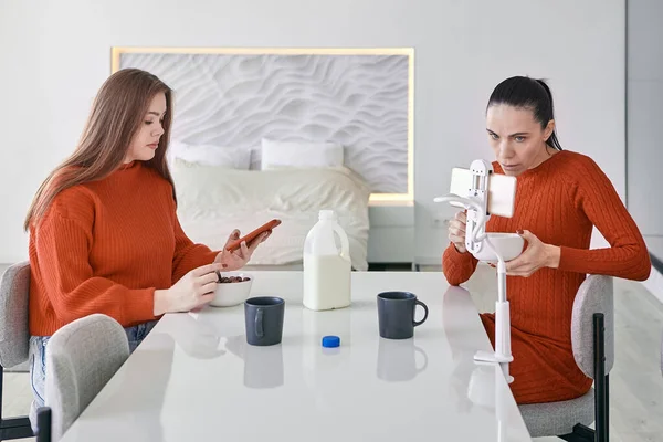 Dos mujeres están desayunando en la mesa de la cocina y buscando información en sus teléfonos inteligentes. —  Fotos de Stock