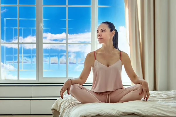 Gran ventana en el dormitorio de la mujer joven sentada en su cama en ropa de casa rosa. — Foto de Stock