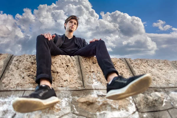 Bottom view of young man in his 20s sitting on stones of pier. — Stock Photo, Image