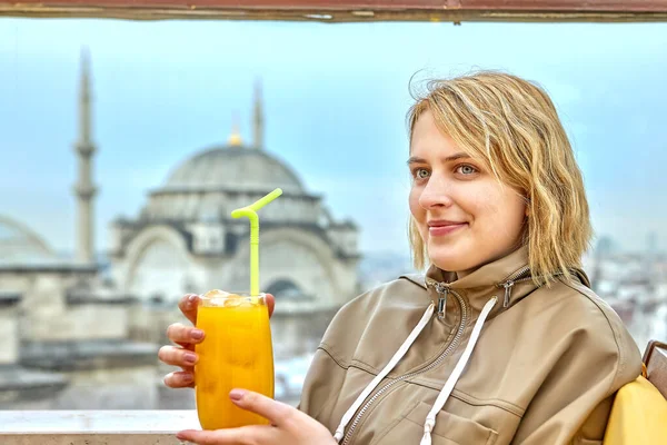 Copo de bebida laranja com gelo e palha nas mãos de uma jovem mulher branca sorridente em um café de Istambul. — Fotografia de Stock