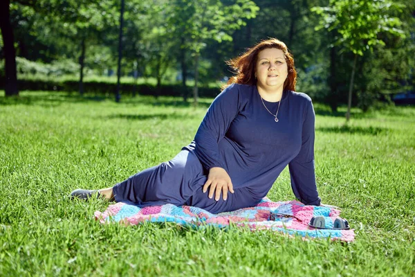Large European woman with pensive expression sits on mat in middle of lawn. — Foto de Stock