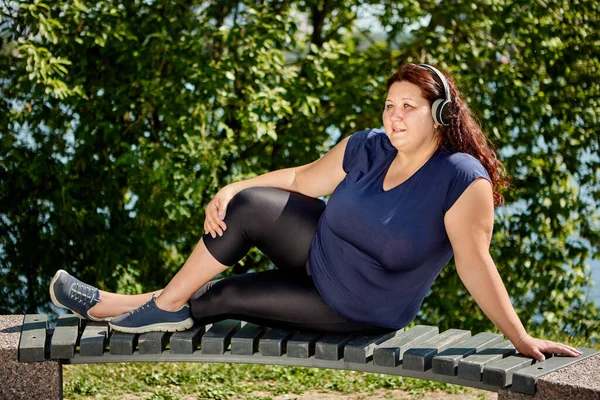 An obese woman with plus size body listens to music through wireless headphones while sitting on bench by river. — Foto de Stock