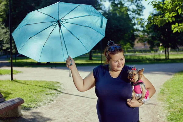 An obese woman covers from sun with an umbrella Yorkshire Terrier, which she holds in her arms. — Stock Photo, Image