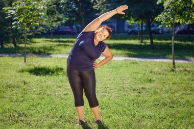 Fat European woman is doing standing side bends exercise on lawn in park. clipart
