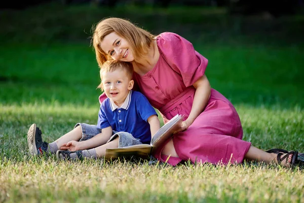 Young smiling woman reads book to little boy sitting on grass in park. — Stock Photo, Image
