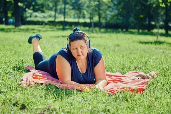 Överviktig kvinna i sladdlösa hörlurar ligger på gräs i parken. — Stockfoto