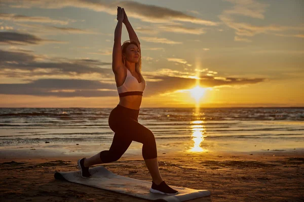 Mujer hace yoga en la estera en la orilla durante el atardecer. — Foto de Stock