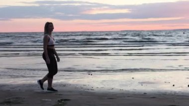Spor giyimli genç beyaz kadın gün batımında kumsalda yürüyor..
