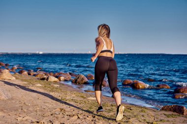 Spor giyimli atletik bir kadın kumlu sahil boyunca koşuyor..