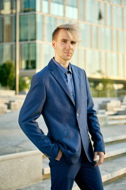 Kendine güvenen genç işadamı ya da mavi takım elbiseli ofis çalışanı açık bir ofis binasının önünde duruyor..