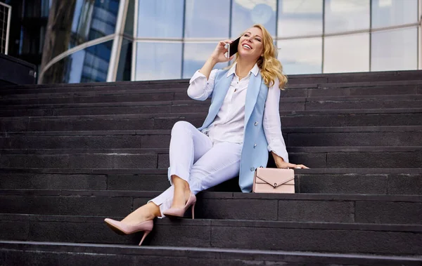 Woman in business suit is talking on phone while sitting on steps in front of an office building. — Stock Photo, Image