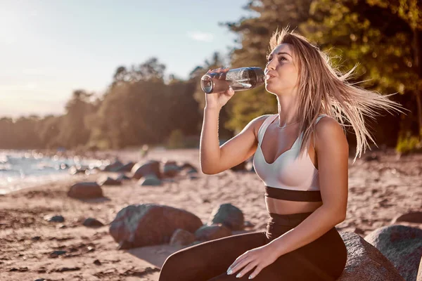 Woman in sportswear drinks water after jogging on sandy sea beach.