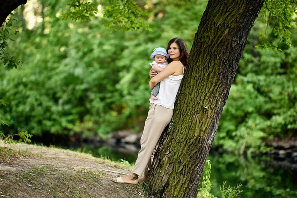 Young woman 26 years with baby boy in her arms walks in city park at summer. — Stock Photo, Image