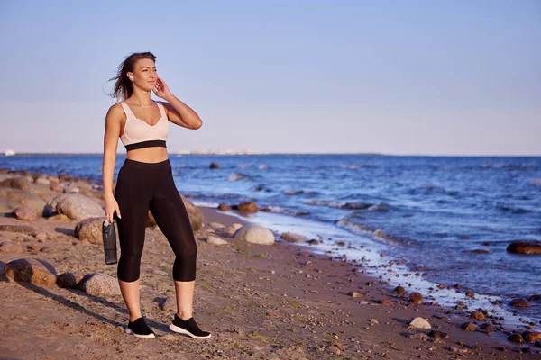 Woman in sportswear stands near seaside with bottle of water during. — Stock Photo, Image