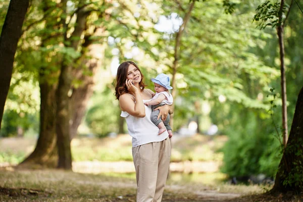 Woman holds little child and talks by cell phone outdoors. — Stock Photo, Image