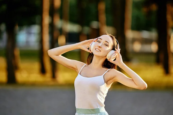 Smiling graceful woman in wireless headphones stands in park. — Stock Photo, Image